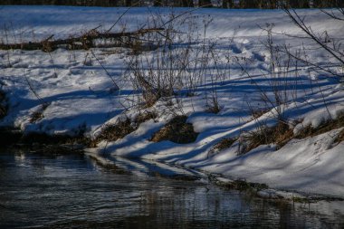 Kışın küçük bir nehir, güneşin ışınları ağaçların arasından geçer. Karlı kıyılar suya yansıyor. Kış manzarası. Kış Masalı. Kış arkaplanı.