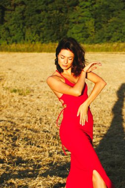 Kırmızı elbiseli güzel bir esmerin portresi biçilmiş buğday tarlasının arka planında, yazın açık havada, gün batımında. Mutlu dans eden kadın