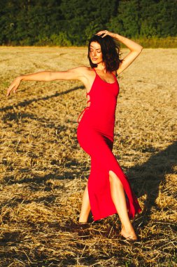 Kırmızı elbiseli güzel bir esmerin portresi biçilmiş buğday tarlasının arka planında, yazın açık havada, gün batımında. Mutlu dans eden kadın