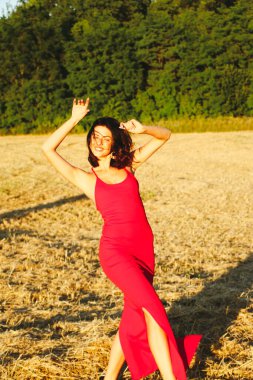 Kırmızı elbiseli güzel bir esmerin portresi biçilmiş buğday tarlasının arka planında, yazın açık havada, gün batımında. Mutlu dans eden kadın