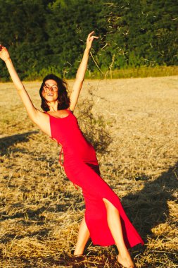 Kırmızı elbiseli güzel bir esmerin portresi biçilmiş buğday tarlasının arka planında, yazın açık havada, gün batımında. Mutlu dans eden kadın