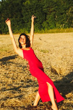 Kırmızı elbiseli güzel bir esmerin portresi biçilmiş buğday tarlasının arka planında, yazın açık havada, gün batımında. Mutlu dans eden kadın