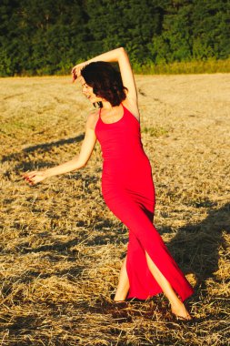 Kırmızı elbiseli güzel bir esmerin portresi biçilmiş buğday tarlasının arka planında, yazın açık havada, gün batımında. Mutlu dans eden kadın