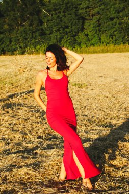 Kırmızı elbiseli güzel bir esmerin portresi biçilmiş buğday tarlasının arka planında, yazın açık havada, gün batımında. Mutlu dans eden kadın