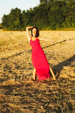 Kırmızı elbiseli güzel bir esmerin portresi biçilmiş buğday tarlasının arka planında, yazın açık havada, gün batımında. Mutlu dans eden kadın