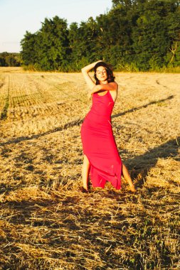 Kırmızı elbiseli güzel bir esmerin portresi biçilmiş buğday tarlasının arka planında, yazın açık havada, gün batımında. Mutlu dans eden kadın