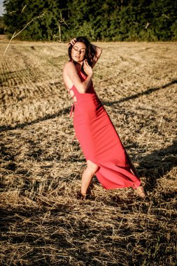 Kırmızı elbiseli güzel bir esmerin portresi biçilmiş buğday tarlasının arka planında, yazın açık havada, gün batımında. Mutlu dans eden kadın