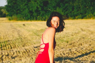 Kırmızı elbiseli güzel bir esmerin portresi biçilmiş buğday tarlasının arka planında, yazın açık havada, gün batımında. Mutlu dans eden kadın