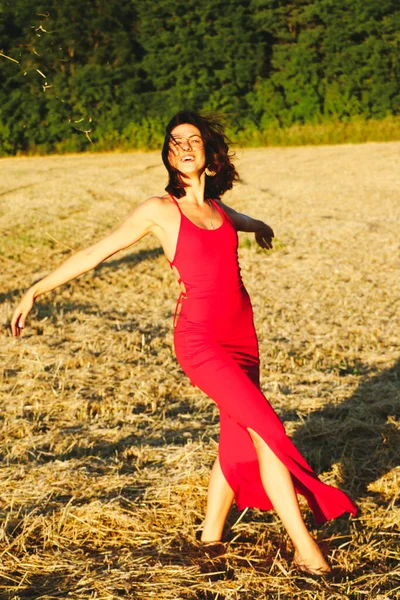 Kırmızı elbiseli güzel bir esmerin portresi biçilmiş buğday tarlasının arka planında, yazın açık havada, gün batımında. Mutlu dans eden kadın