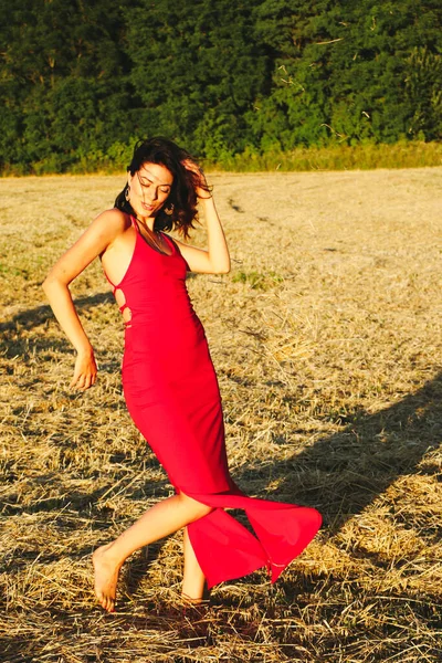 Kırmızı elbiseli güzel bir esmerin portresi biçilmiş buğday tarlasının arka planında, yazın açık havada, gün batımında. Mutlu dans eden kadın