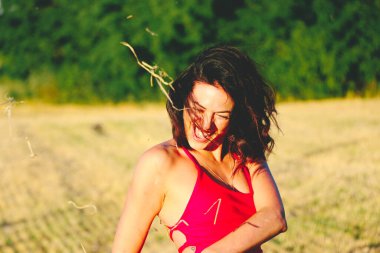 Kırmızı elbiseli güzel bir esmerin portresi biçilmiş buğday tarlasının arka planında, yazın açık havada, gün batımında. Mutlu dans eden kadın