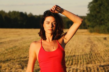 Kırmızı elbiseli güzel bir esmerin portresi biçilmiş buğday tarlasının arka planında, yazın açık havada, gün batımında. Mutlu dans eden kadın