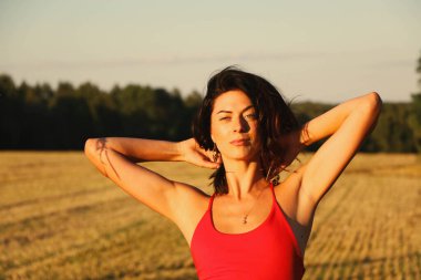 Kırmızı elbiseli güzel bir esmerin portresi biçilmiş buğday tarlasının arka planında, yazın açık havada, gün batımında. Mutlu dans eden kadın