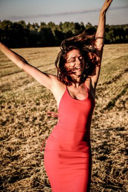 Kırmızı elbiseli güzel bir esmerin portresi biçilmiş buğday tarlasının arka planında, yazın açık havada, gün batımında. Mutlu dans eden kadın