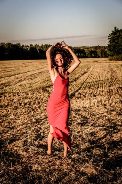 Kırmızı elbiseli güzel bir esmerin portresi biçilmiş buğday tarlasının arka planında, yazın açık havada, gün batımında. Mutlu dans eden kadın
