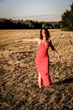 Kırmızı elbiseli güzel bir esmerin portresi biçilmiş buğday tarlasının arka planında, yazın açık havada, gün batımında. Mutlu dans eden kadın