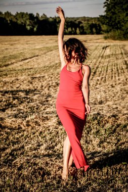 Kırmızı elbiseli güzel bir esmerin portresi biçilmiş buğday tarlasının arka planında, yazın açık havada, gün batımında. Mutlu dans eden kadın