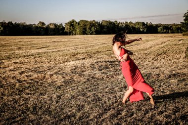 Kırmızı elbiseli güzel bir esmerin portresi biçilmiş buğday tarlasının arka planında, yazın açık havada, gün batımında. Mutlu dans eden kadın