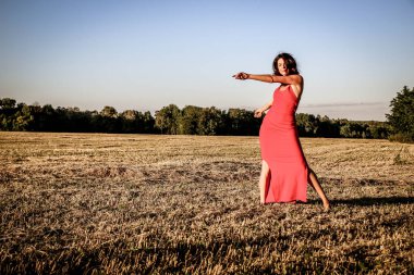 Kırmızı elbiseli güzel bir esmerin portresi biçilmiş buğday tarlasının arka planında, yazın açık havada, gün batımında. Mutlu dans eden kadın