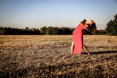 Kırmızı elbiseli güzel bir esmerin portresi biçilmiş buğday tarlasının arka planında, yazın açık havada, gün batımında. Mutlu dans eden kadın