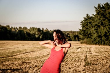 Kırmızı elbiseli güzel bir esmerin portresi biçilmiş buğday tarlasının arka planında, yazın açık havada, gün batımında. Mutlu dans eden kadın