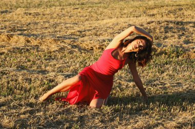 Kırmızı elbiseli güzel bir esmerin portresi biçilmiş buğday tarlasının arka planında, yazın açık havada, gün batımında. Mutlu dans eden kadın