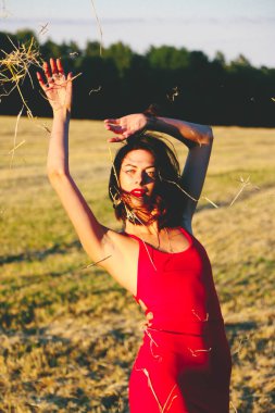 Kırmızı elbiseli güzel bir esmerin portresi biçilmiş buğday tarlasının arka planında, yazın açık havada, gün batımında. Mutlu dans eden kadın