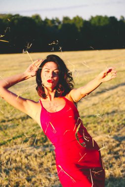 Kırmızı elbiseli güzel bir esmerin portresi biçilmiş buğday tarlasının arka planında, yazın açık havada, gün batımında. Mutlu dans eden kadın