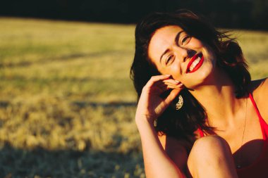 Kırmızı elbiseli güzel bir esmerin portresi biçilmiş buğday tarlasının arka planında, yazın açık havada, gün batımında. Oturan kadın