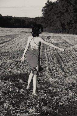 Yazın açık havada, gün batımında, biçilmiş buğday tarlasının arka planında, elbisenin içinde genç ve güzel bir esmerin portresi. Mutlu dans eden kadın, siyah beyaz fotoğraf