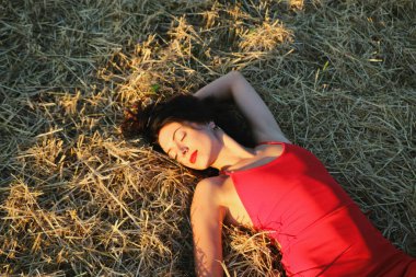 Parlak dudaklı, kırmızı elbiseli, güzel bir esmer kadın samanlıkta yatıyor. Yaz akşamı gün batımı ışığı. Üst görünüm yakın plan