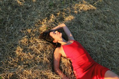 Parlak dudaklı, kırmızı elbiseli, güzel bir esmer kadın samanlıkta yatıyor. Yaz akşamı gün batımı ışığı. Üst görünüm yakın plan