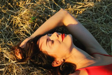 Parlak dudaklı, kırmızı elbiseli, güzel bir esmer kadın samanlıkta yatıyor. Yaz akşamı gün batımı ışığı. Üst görünüm yakın plan