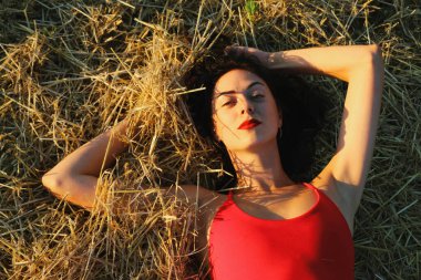 Parlak dudaklı, kırmızı elbiseli, güzel bir esmer kadın samanlıkta yatıyor. Yaz akşamı gün batımı ışığı. Üst görünüm yakın plan