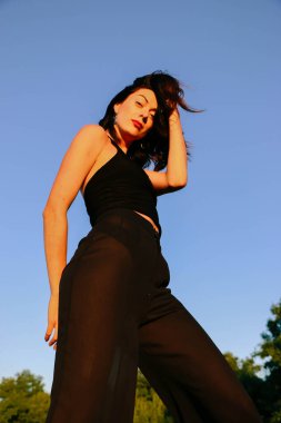 Güneşli bir yaz akşamında, gün batımında poz veren şık siyah elbiseli güzel bir esmer. Çekici kadın mutlu bir şekilde dans ediyor, mavi gökyüzüne karşı poz veriyor.