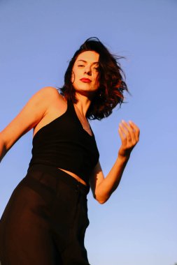 Güneşli bir yaz akşamında, gün batımında poz veren şık siyah elbiseli güzel bir esmer. Çekici kadın mutlu bir şekilde dans ediyor, mavi gökyüzüne karşı poz veriyor.