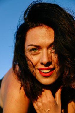 Güneşli bir yaz akşamında, gün batımında poz veren şık siyah elbiseli güzel bir esmer. Çekici bir kadın mutlu bir şekilde dans ediyor, mavi gökyüzüne karşı yakın plan pozlar veriyor.