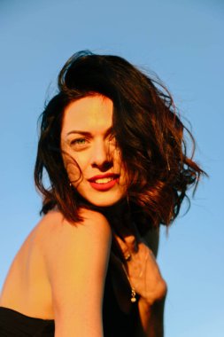 Güneşli bir yaz akşamında, gün batımında poz veren şık siyah elbiseli güzel bir esmer. Çekici bir kadın mutlu bir şekilde dans ediyor, mavi gökyüzüne karşı yakın plan pozlar veriyor.
