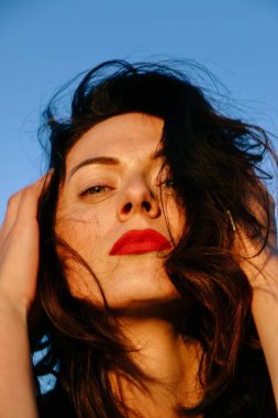 Güneşli bir yaz akşamında, gün batımında poz veren şık siyah elbiseli güzel bir esmer. Çekici bir kadın mutlu bir şekilde dans ediyor, mavi gökyüzüne karşı yakın plan pozlar veriyor.