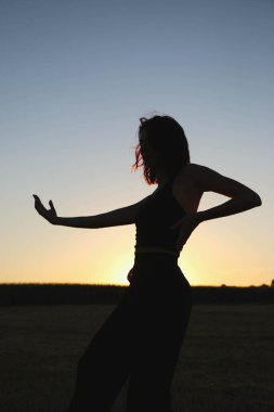 Güneşli bir yaz akşamında, gün batımında poz veren şık siyah elbiseli güzel bir esmer. Çekici kadın mutlu bir şekilde dans ediyor, mavi gökyüzüne karşı poz veriyor.