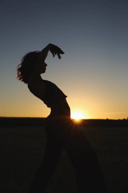 Güneşli bir yaz akşamında, gün batımında poz veren şık siyah elbiseli güzel bir esmer. Çekici kadın mutlu bir şekilde dans ediyor, mavi gökyüzüne karşı poz veriyor.