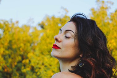 Güneşli bir yaz akşamında şık siyah elbiseli genç ve güzel bir esmer. Çekici kadın mutlu bir şekilde dans ediyor, yeşil ağaçların ve mavi gökyüzünün arka planında poz veriyor.