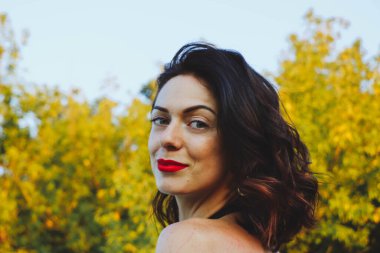 Güneşli bir yaz akşamında şık siyah elbiseli genç ve güzel bir esmer. Çekici kadın mutlu bir şekilde dans ediyor, yeşil ağaçların ve mavi gökyüzünün arka planında poz veriyor.