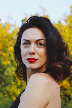 Güneşli bir yaz akşamında şık siyah elbiseli genç ve güzel bir esmer. Çekici kadın mutlu bir şekilde dans ediyor, yeşil ağaçların ve mavi gökyüzünün arka planında poz veriyor.