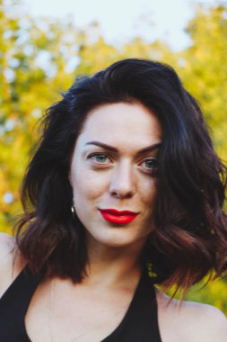 Güneşli bir yaz akşamında şık siyah elbiseli genç ve güzel bir esmer. Çekici kadın mutlu bir şekilde dans ediyor, yeşil ağaçların ve mavi gökyüzünün arka planında poz veriyor.