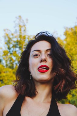 Güneşli bir yaz akşamında şık siyah elbiseli genç ve güzel bir esmer. Çekici kadın mutlu bir şekilde dans ediyor, yeşil ağaçların ve mavi gökyüzünün arka planında poz veriyor.