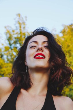 Güneşli bir yaz akşamında şık siyah elbiseli genç ve güzel bir esmer. Çekici kadın mutlu bir şekilde dans ediyor, yeşil ağaçların ve mavi gökyüzünün arka planında poz veriyor.