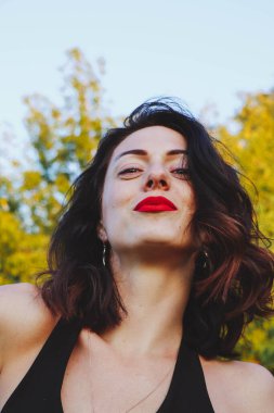 Güneşli bir yaz akşamında şık siyah elbiseli genç ve güzel bir esmer. Çekici kadın mutlu bir şekilde dans ediyor, yeşil ağaçların ve mavi gökyüzünün arka planında poz veriyor.