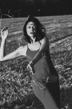 Yazın açık havada, gün batımında, biçilmiş buğday tarlasının arka planında, elbisenin içinde genç ve güzel bir esmerin portresi. Mutlu dans eden kadın, siyah beyaz fotoğraf