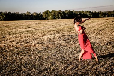 Kırmızı elbiseli güzel bir esmerin portresi biçilmiş buğday tarlasının arka planında, yazın açık havada, gün batımında. Mutlu dans eden kadın